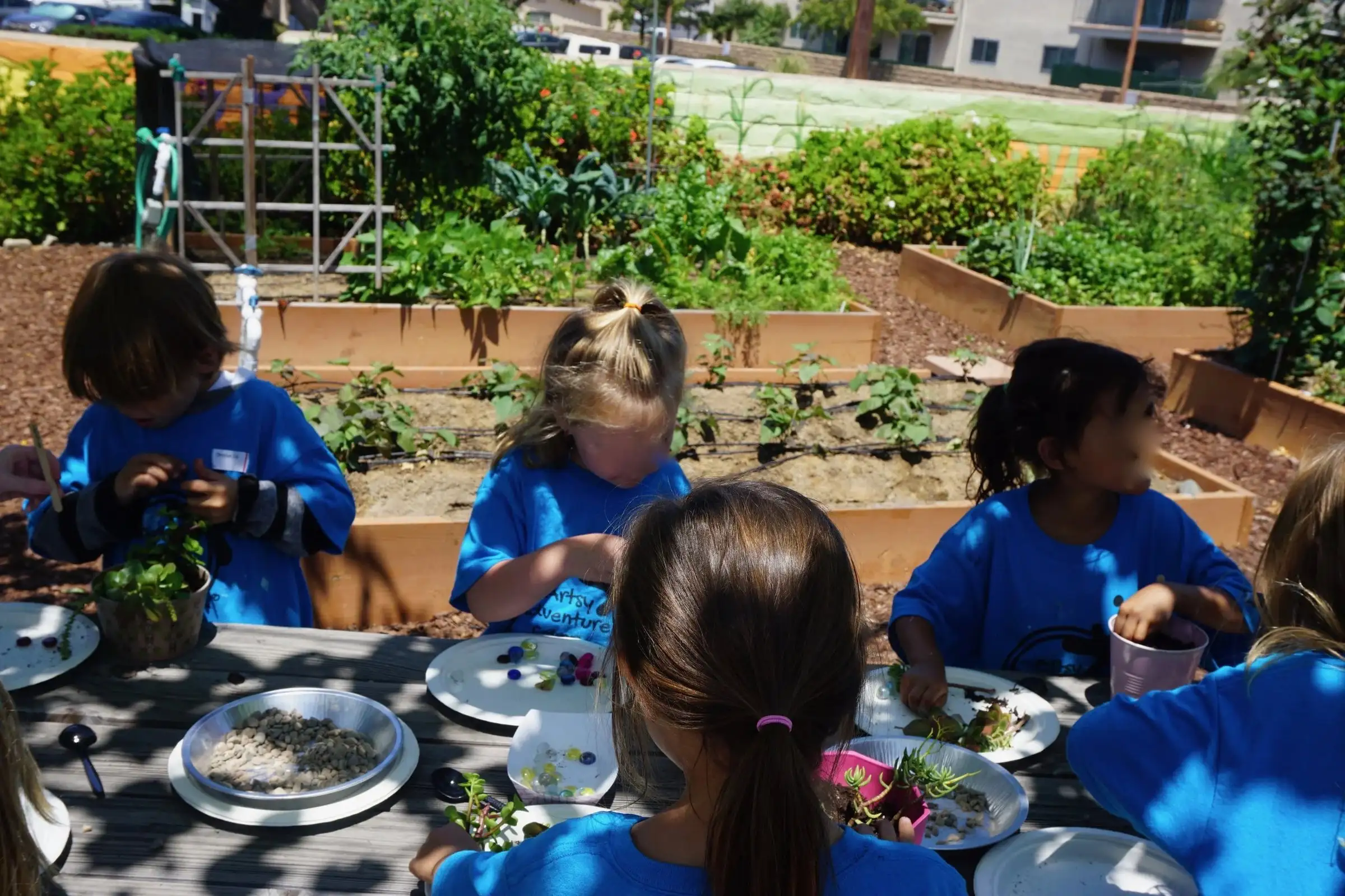 Children in camp shirts eating lunch together at a table beside the garden beds