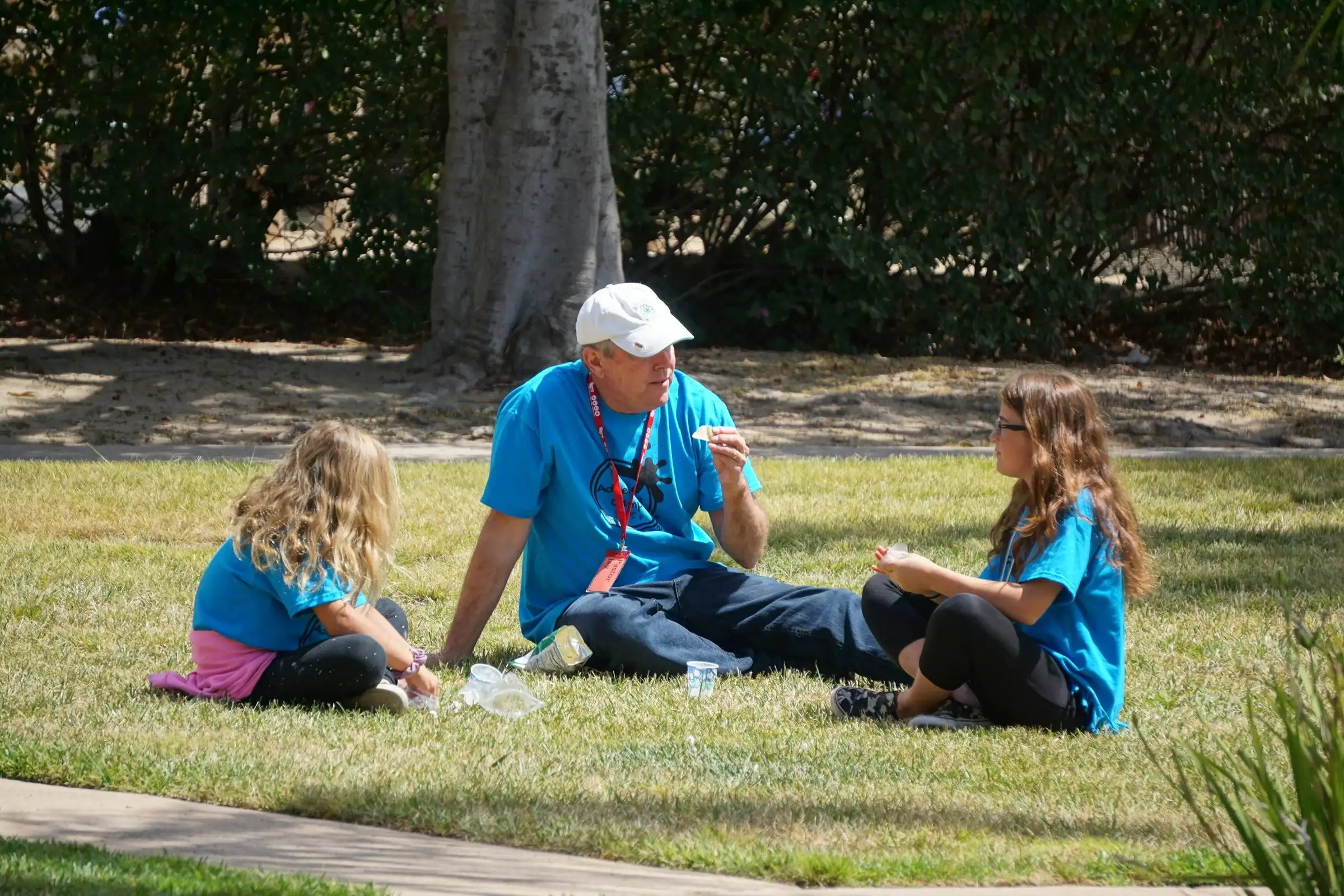 A volunteer in a baseball cap sits on the lawn talking with two girls during summer camp