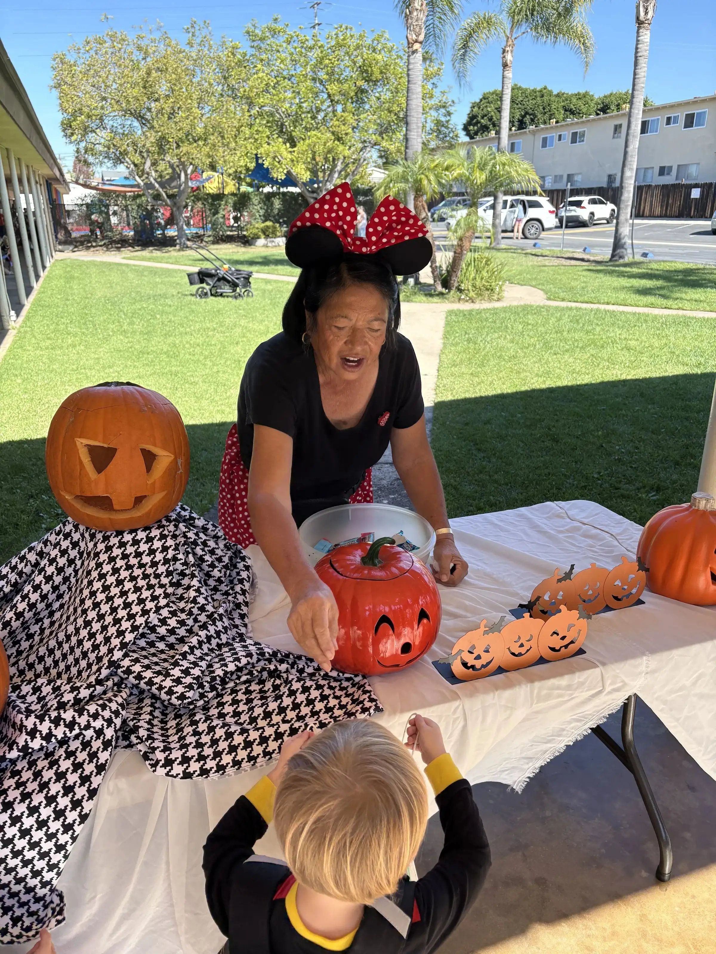 A church volunteer in Minnie Mouse ears helping a young child decorate pumpkins on the lawn