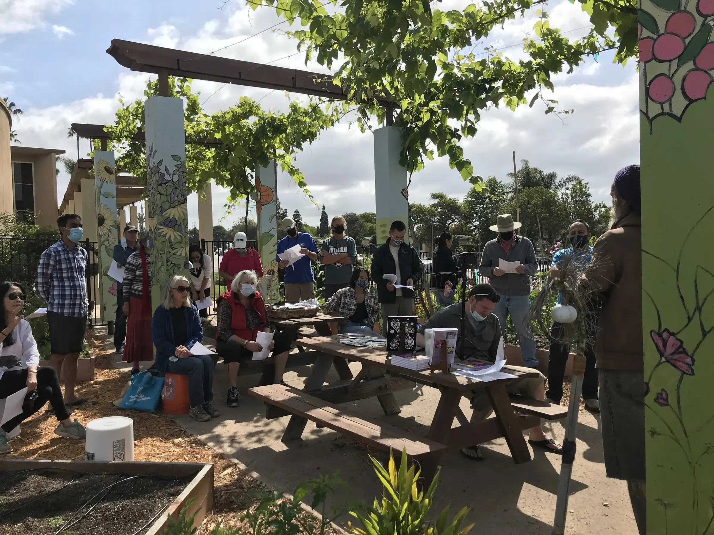 Gardeners gathered at picnic tables beneath a vine-covered arbor in the community garden